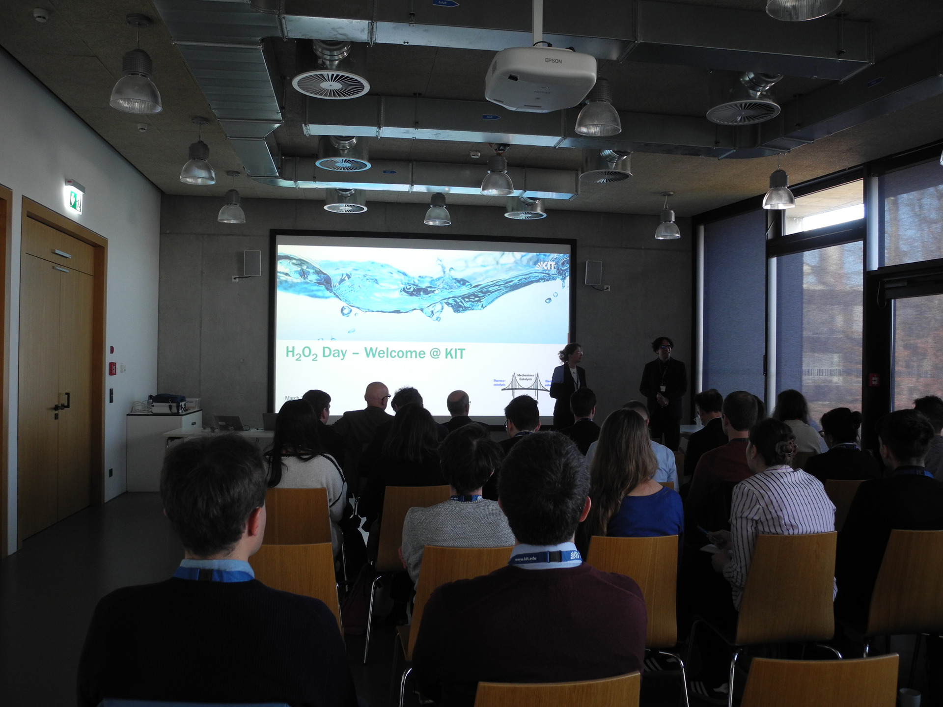 Vortrag im Konferenzraum: Pr&auml;sentation auf Leinwand mit Wasser-Symbol, Zuh&ouml;rer sitzen in Stuhlreihen.