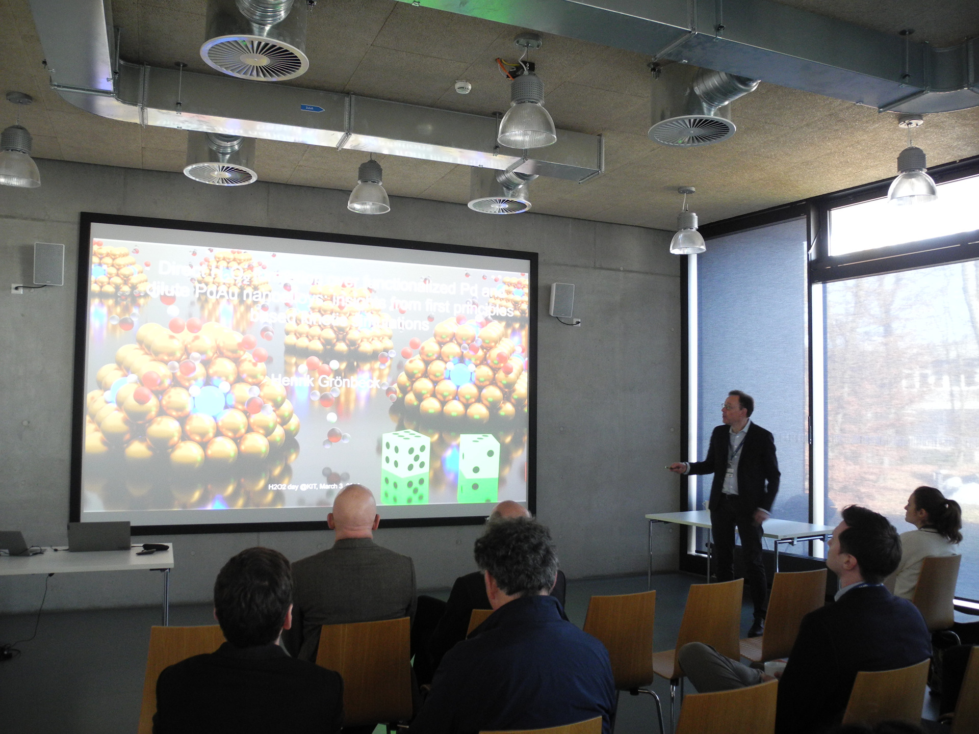 Vortrag in modernem Konferenzraum: Pr&auml;sentationsfolie mit bunten Kugeln, W&uuml;rfeln und Diagrammen.