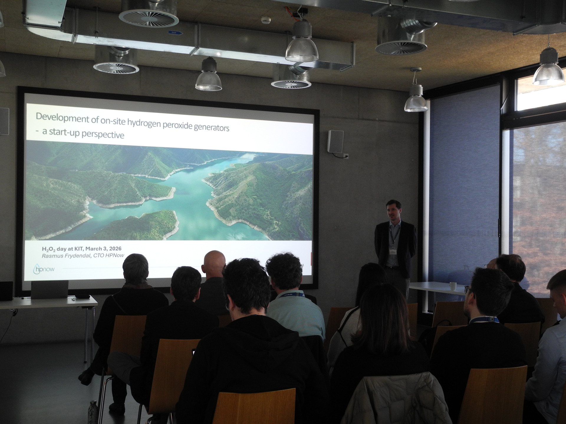 Vortrag vor Publikum in Konferenzraum; Pr&auml;sentationsfolie mit Landschaftsbild (Staudamm-/Stausee).