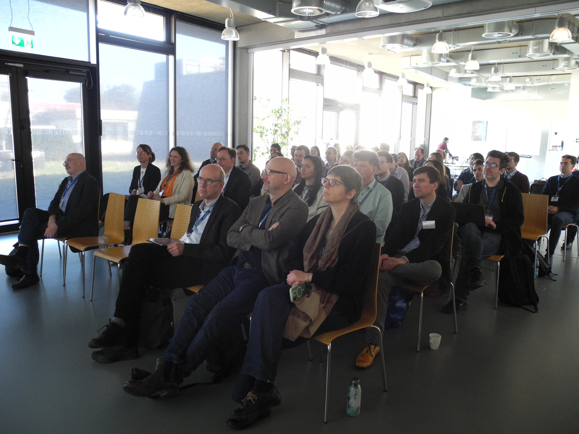 Konferenzsaal mit circa 25 Menschen, sitzen in Reihen und schauen aus Richtung B&uuml;hne/Leinwand.