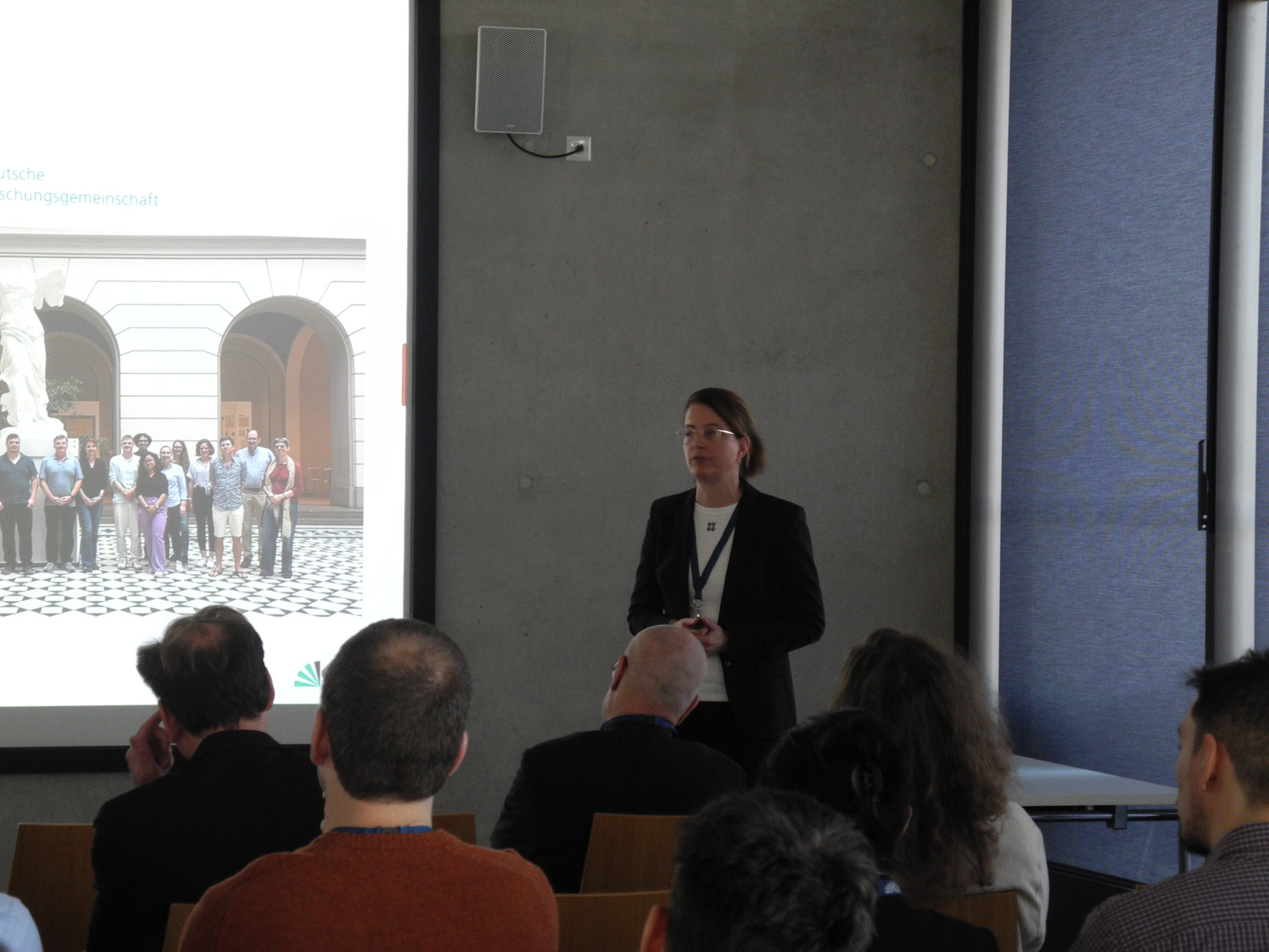 Gruppe von Menschen vor einer Projektion mit einer redenden Frau an der Wand.