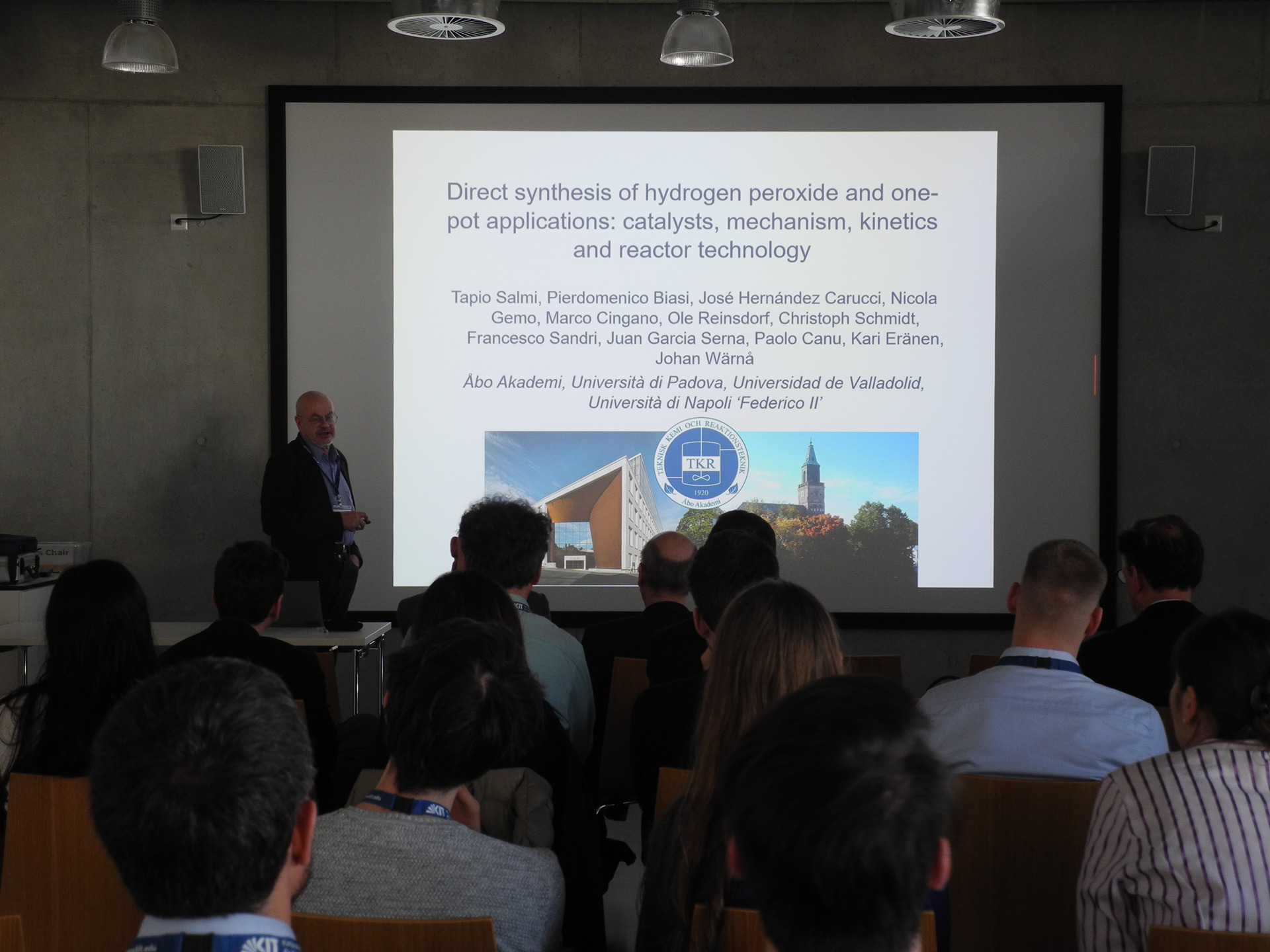 Vortragende vor einer projetierten Folie in einem H&ouml;rsaal w&auml;hrend Konferenz, Publikum schaut zu.