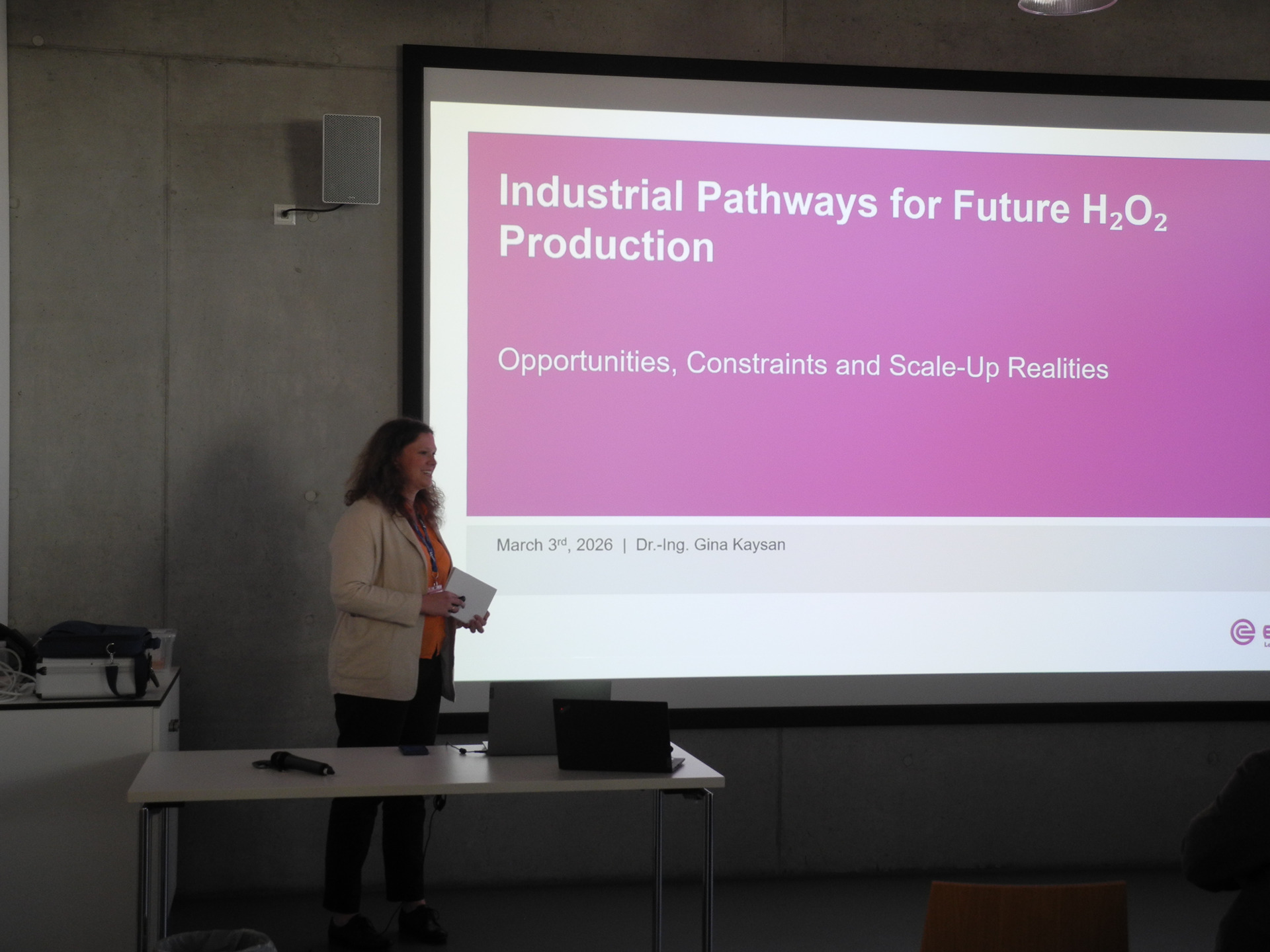 Vortragende/r steht neben Leinwand mit Folie „Industrial Pathways for Future H2O2 Production“ im Konferenzraum.