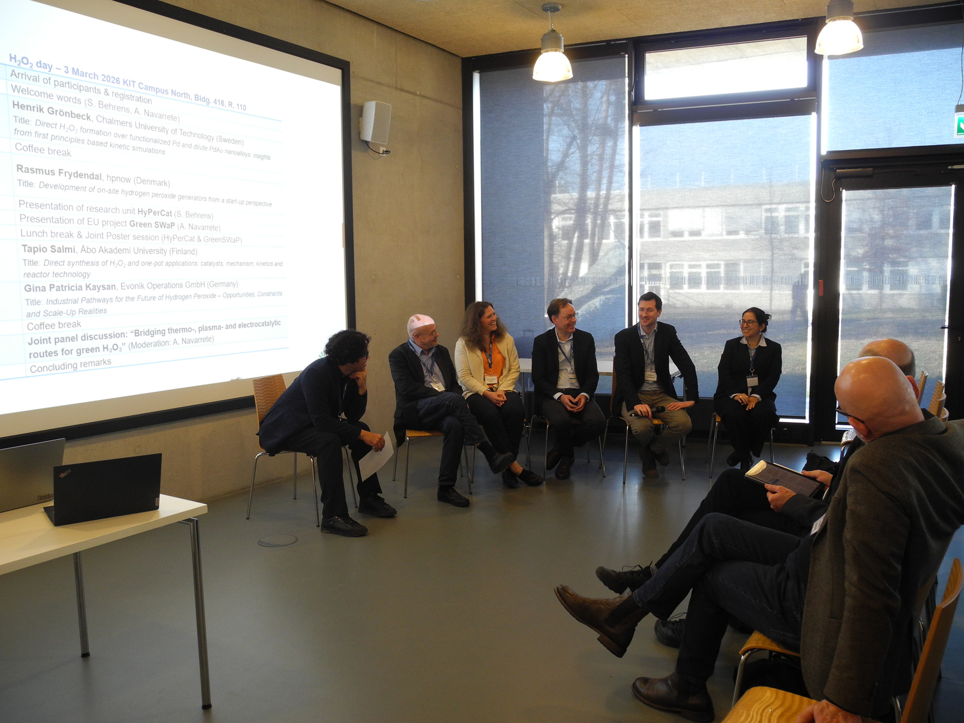 Gruppe von Menschen sitzt in einem H&ouml;rsaal in einem Panel- oder Workshop-Szenario, Speaker am Vortrag, Pr&auml;sentationsfolie rechts sichtbar.